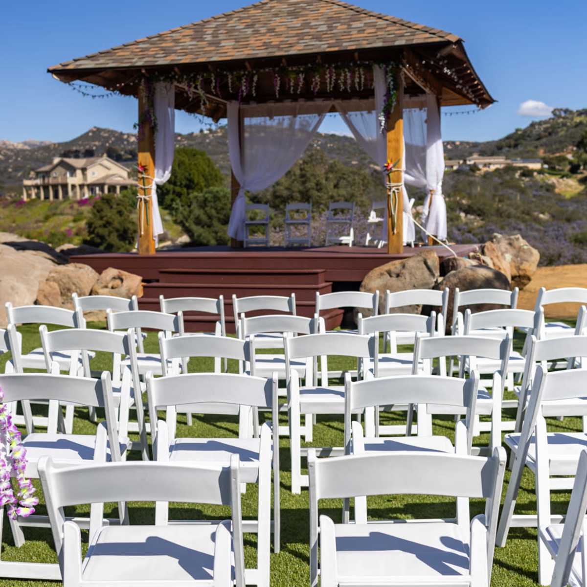 A gazebo decorated with drapes and flowers stands on a grassy area facing rows of white chairs set up for an outdoor event.
