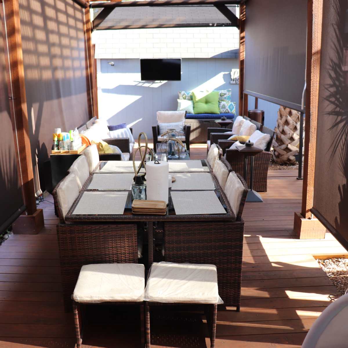 A shaded outdoor seating area featuring a large dining table surrounded by multiple chairs, with a small couch and decorative pillows in the background.