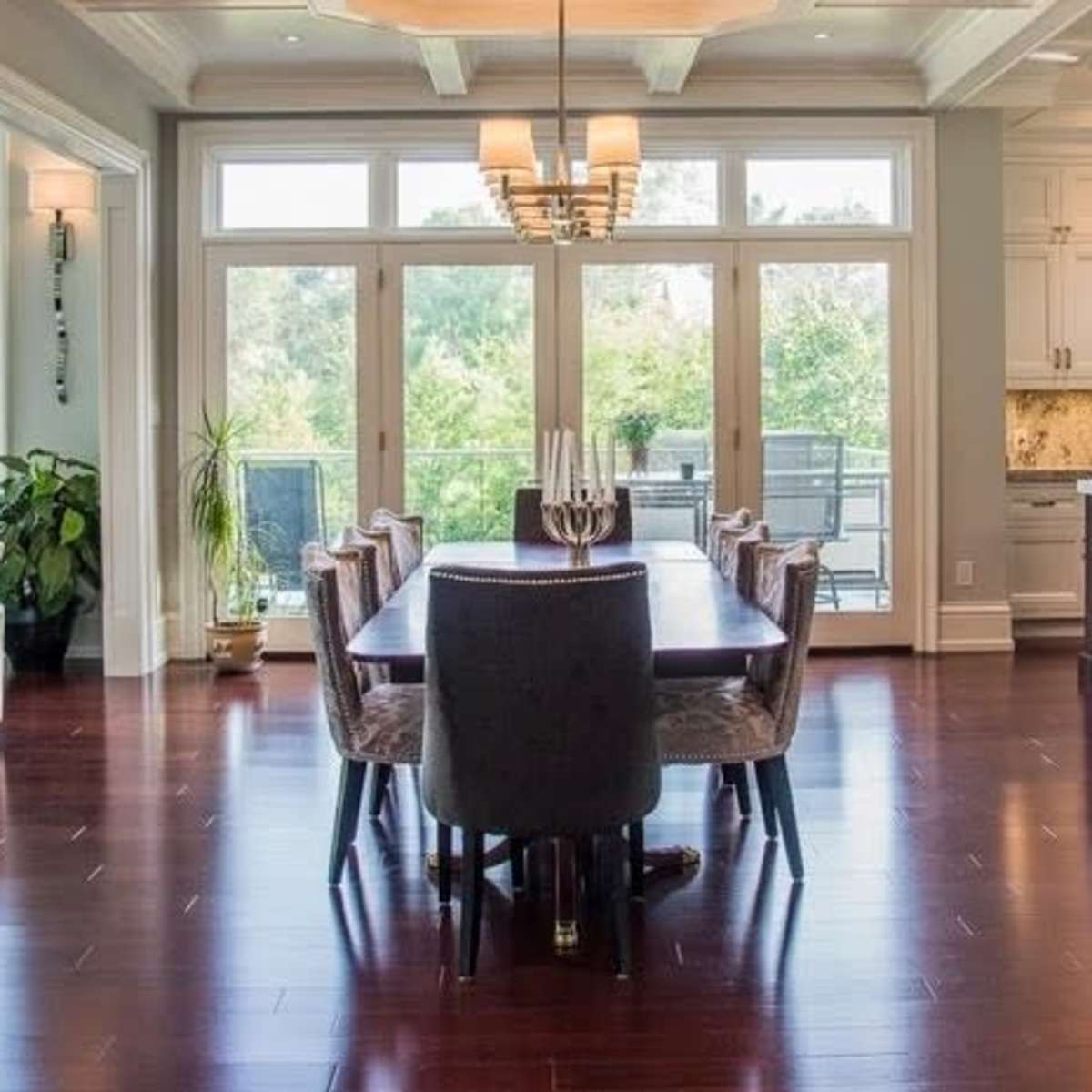 A spacious dining area with a long table surrounded by chairs, adjacent to a modern kitchen with large windows offering a view of greenery outside.