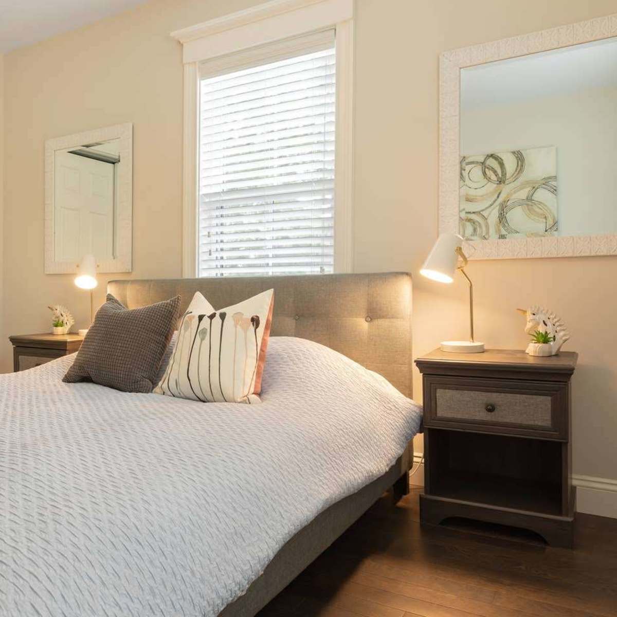 A bedroom with a gray upholstered bed, two bedside tables, a mirror, and a window with blinds.