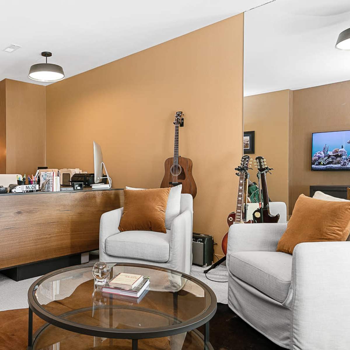 A modern office space featuring a wooden desk, two light-colored chairs, a round glass coffee table, and musical instruments displayed on a wall.