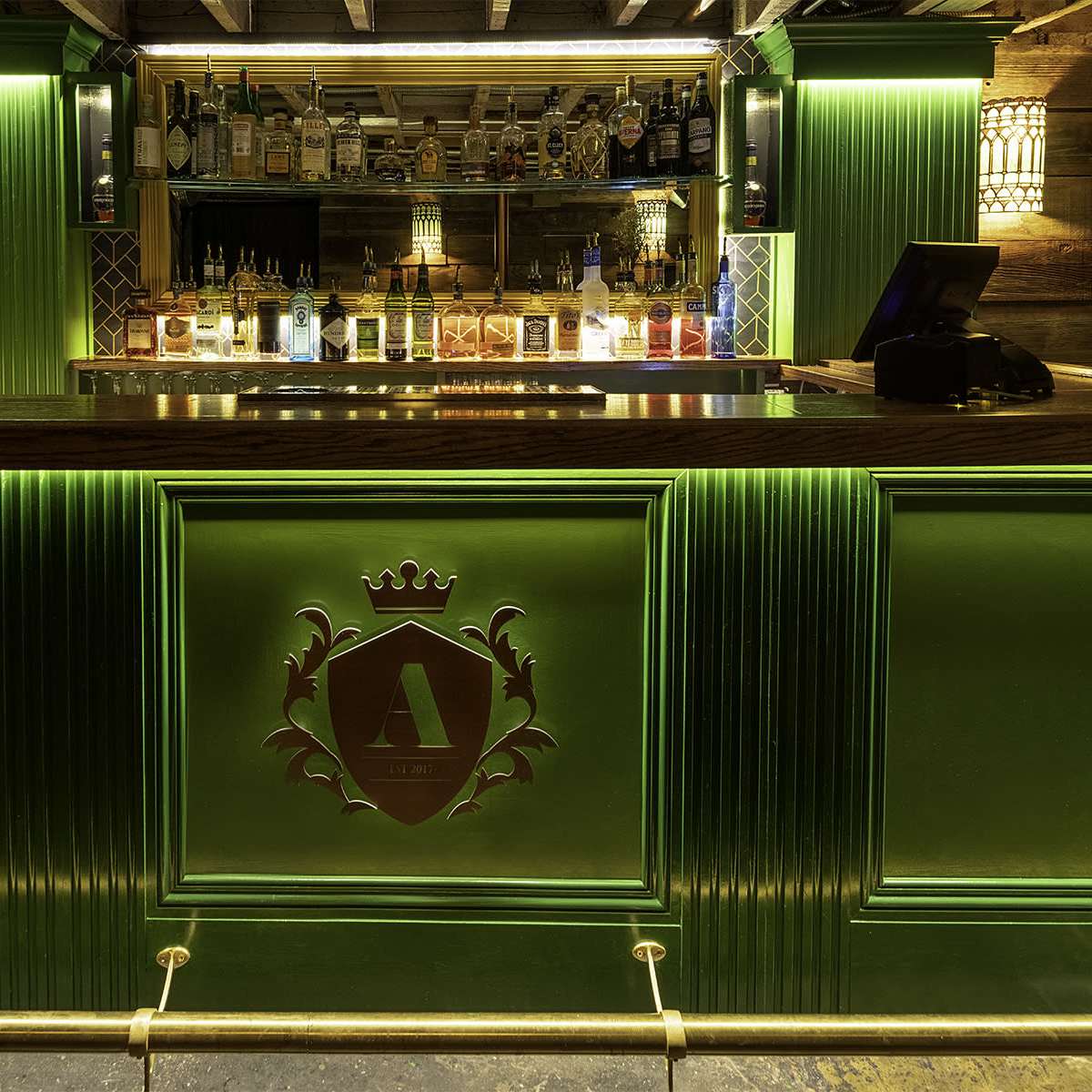 A bar counter with a wooden surface and green illuminated panels, featuring various bottles of liquor displayed on shelves behind it.