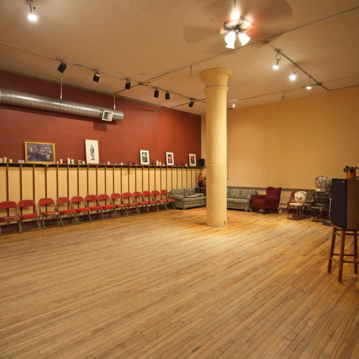 An empty room with wooden flooring, a central column, red and beige walls, and several chairs arranged along one side.