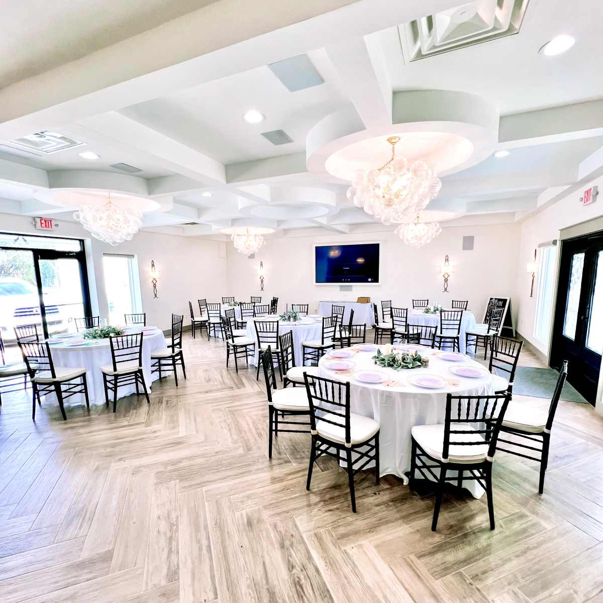 A spacious event room features multiple round tables set for dining and decorated with white tablecloths, alongside elegant chandeliers and a large screen on the wall.