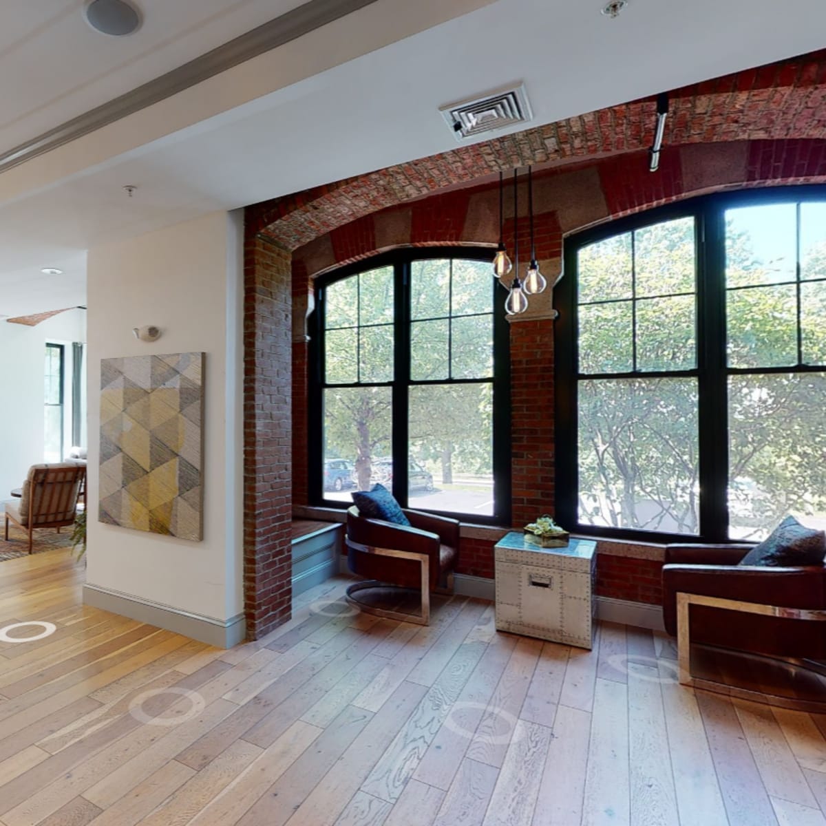 A brightly lit room features large windows, hardwood floors, and various seating areas, including two chairs and artwork on the walls.