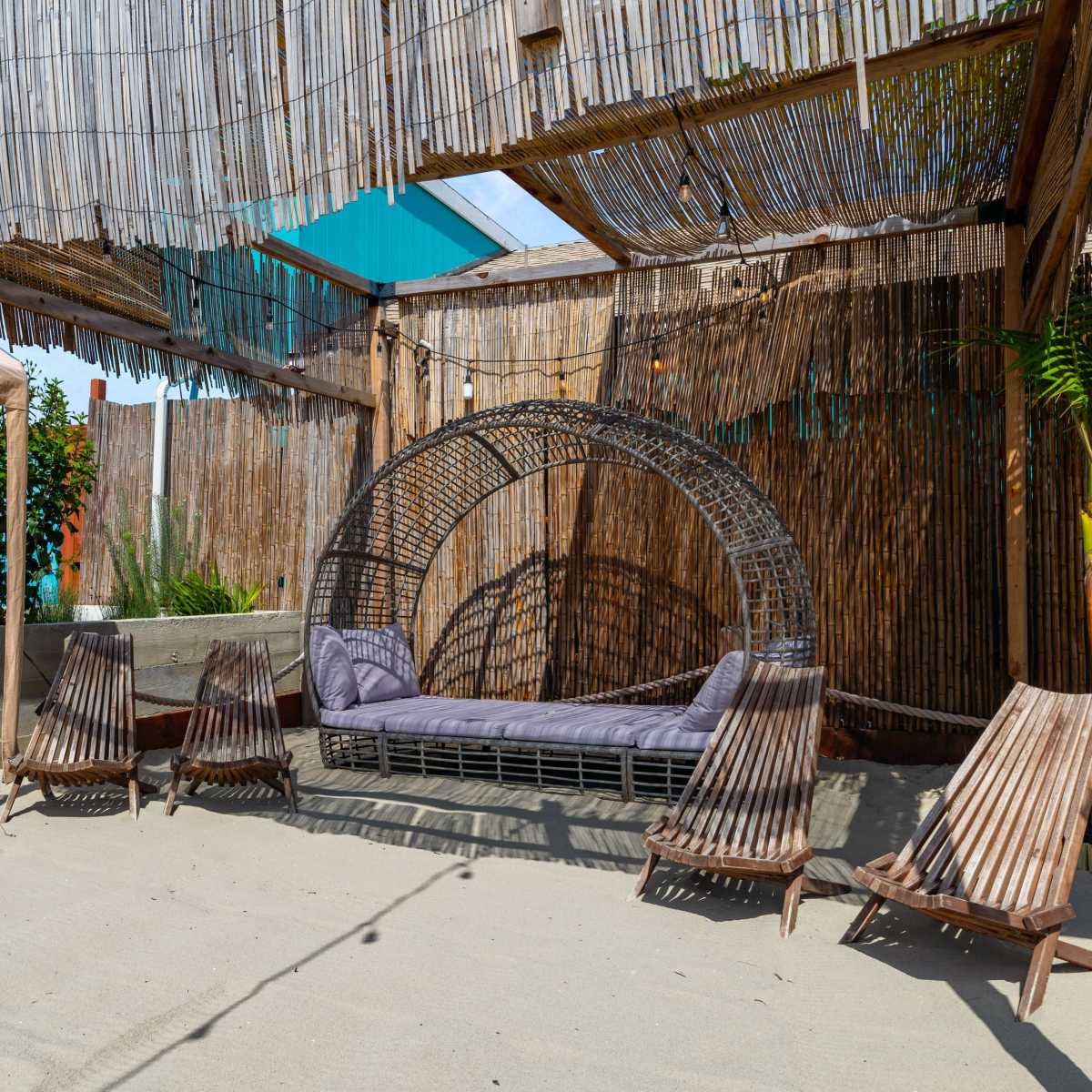 A cozy seating area with a circular wicker sofa and four wooden lounge chairs is set in a sandy space partially shaded by a bamboo structure.