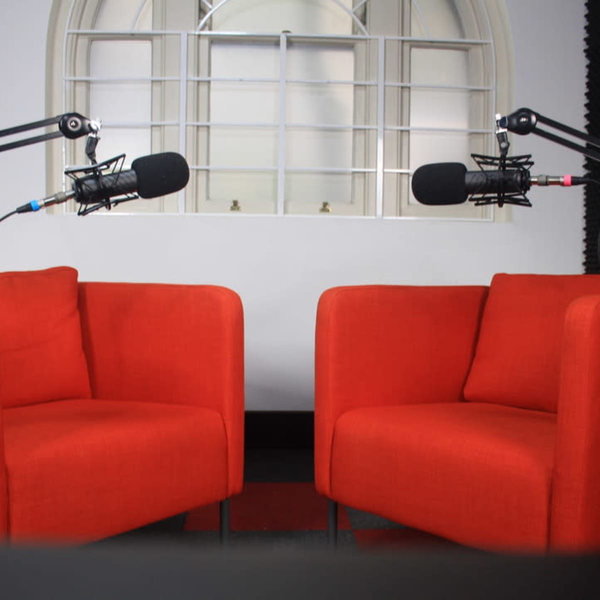Two red armchairs positioned facing each other, each equipped with a microphone on an adjustable stand, set against a wall with a large window.