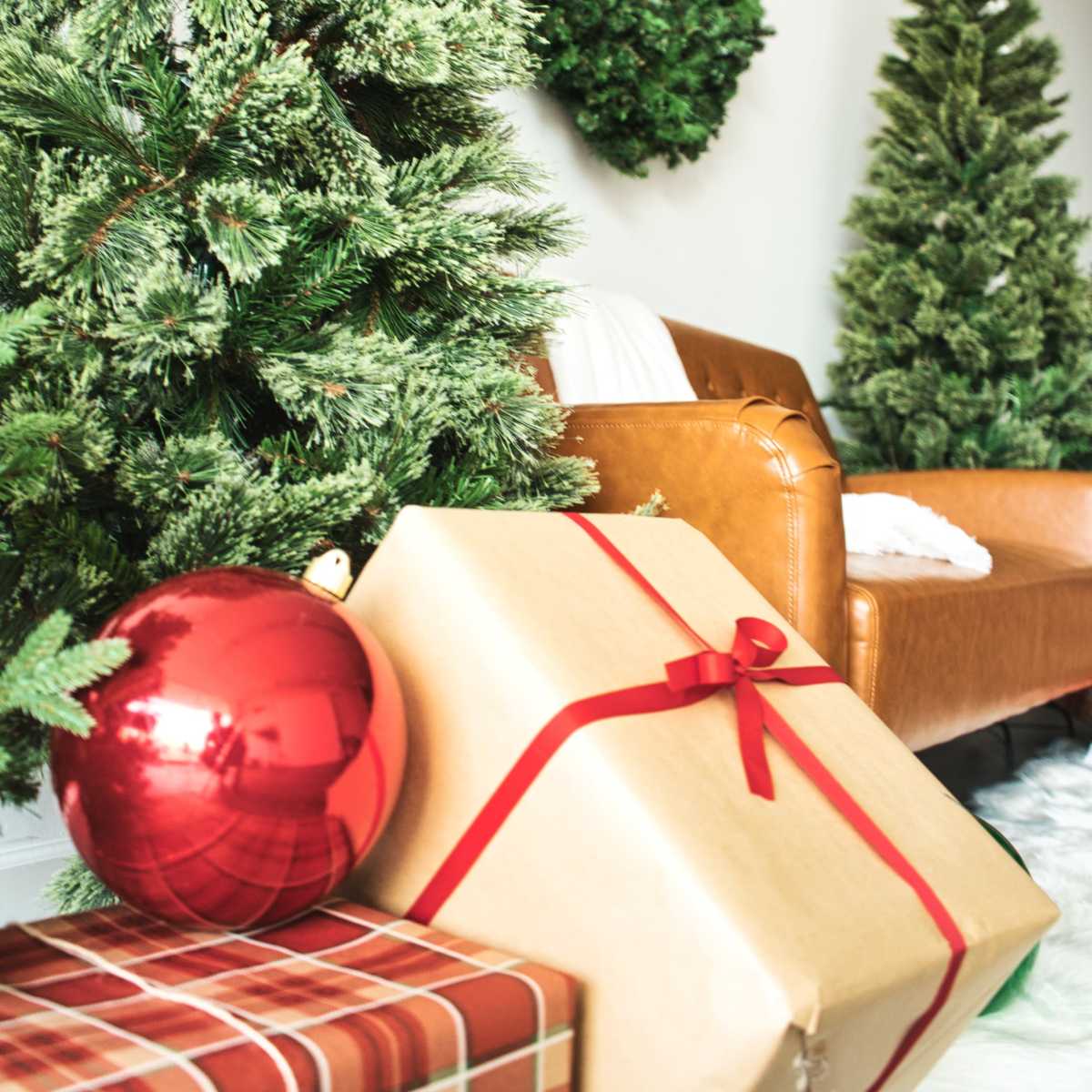 A cozy living room decorated with two Christmas trees, a wreath on the wall, and stacked gifts beside a brown leather chair.