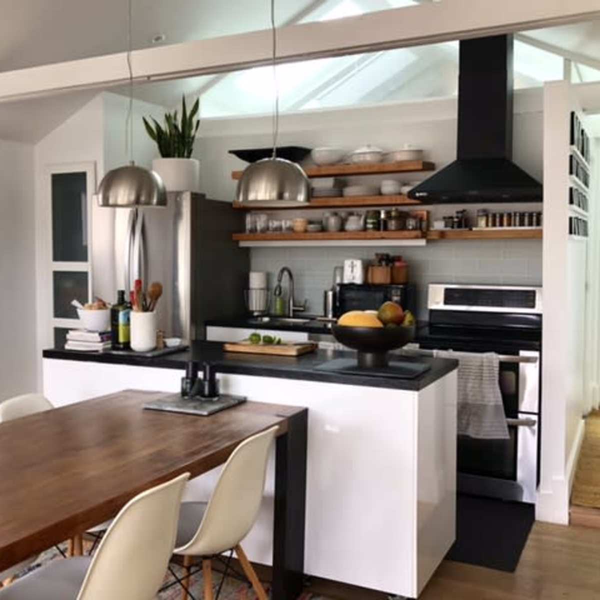 A modern kitchen featuring a central wooden dining table, sleek cabinetry, open shelving with kitchenware, and stainless steel appliances.