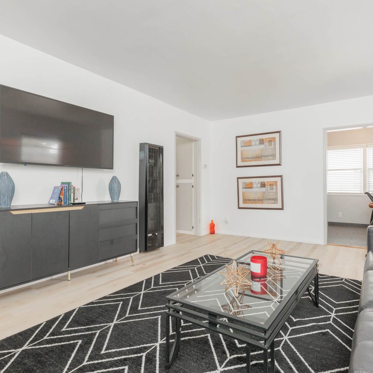 A modern living room with a dark sofa, a glass coffee table, a wall-mounted television, and framed artwork on the walls.