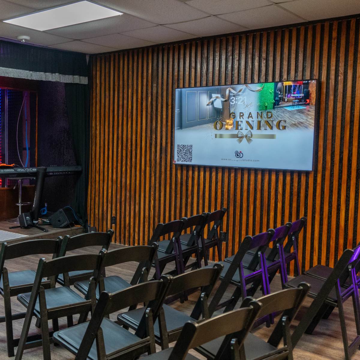 A small performance space with a stage featuring musical instruments, a microphone, and rows of chairs facing the stage, along with a large screen announcing a grand opening.