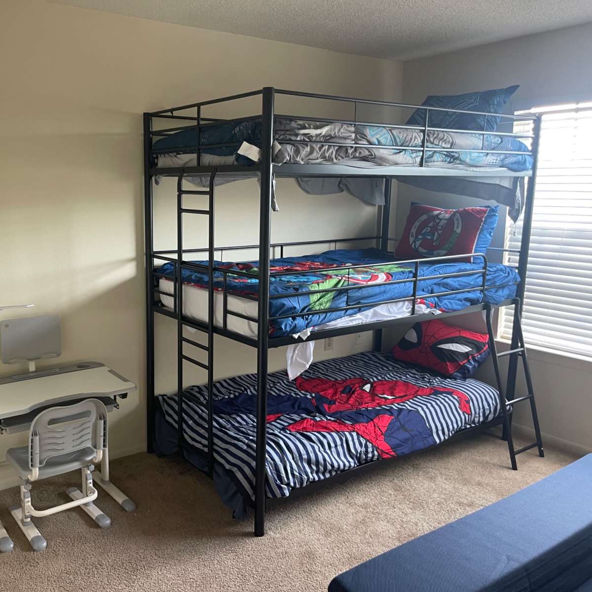 A metal bunk bed with superhero-themed bedding is positioned next to a small desk and chair in a room with beige walls and a window.