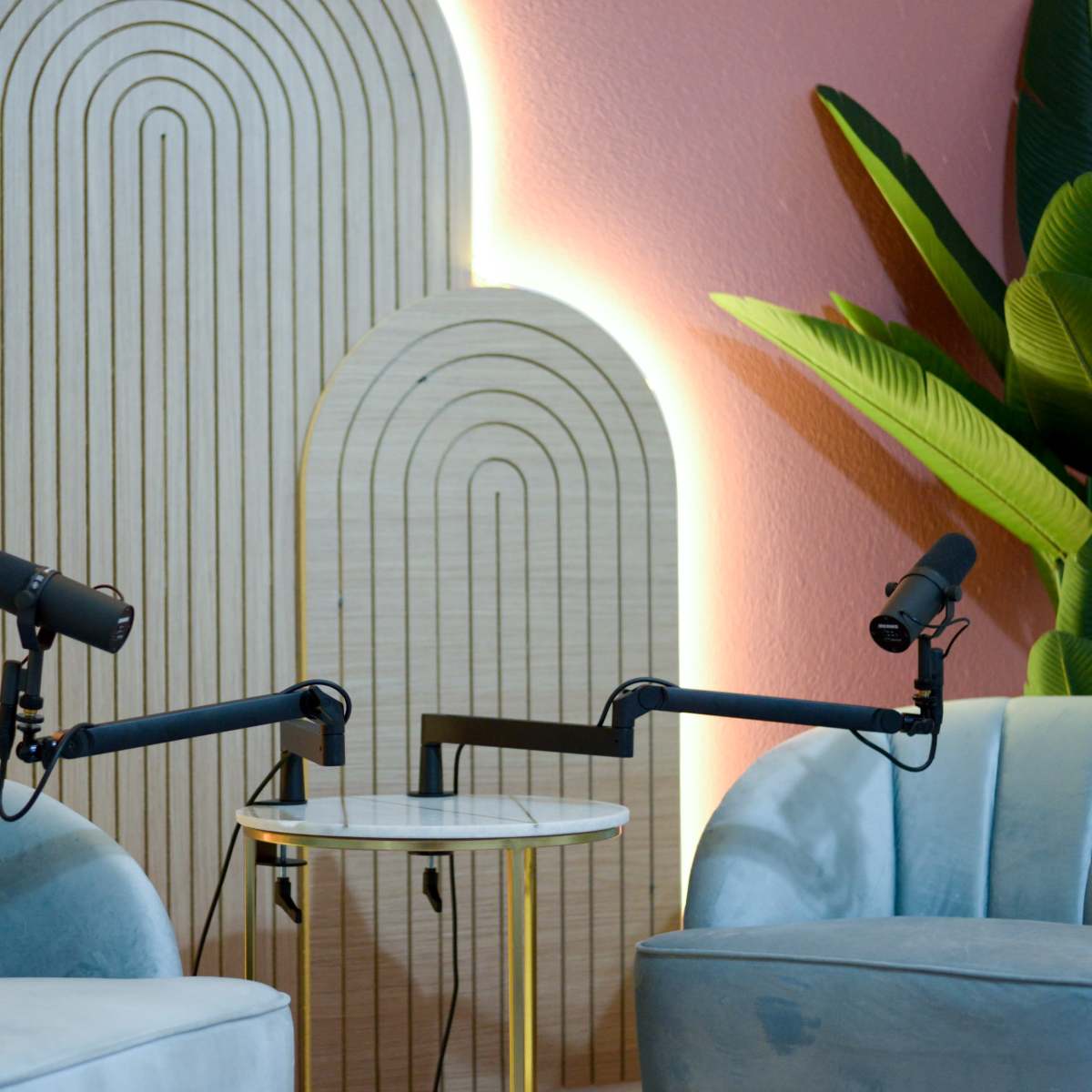 Two microphones positioned on adjustable arms facing two blue velvet chairs beside a round table, set against a backdrop of wooden arch designs and green plants.