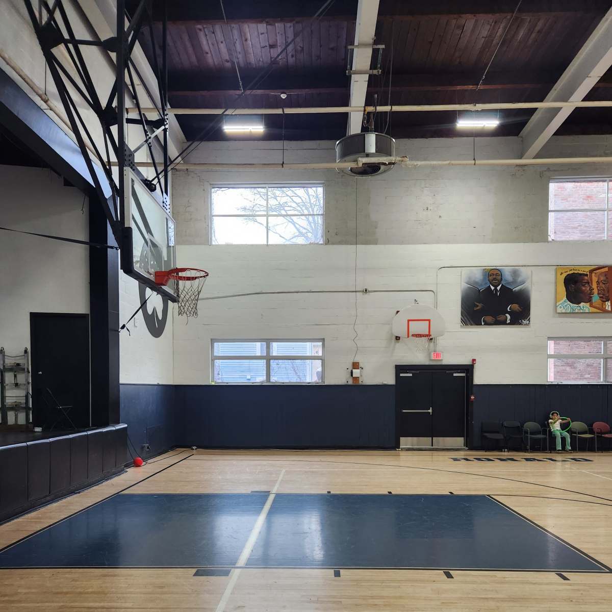 A basketball court featuring a hoop, hardwood flooring, and large windows alongside wall murals.