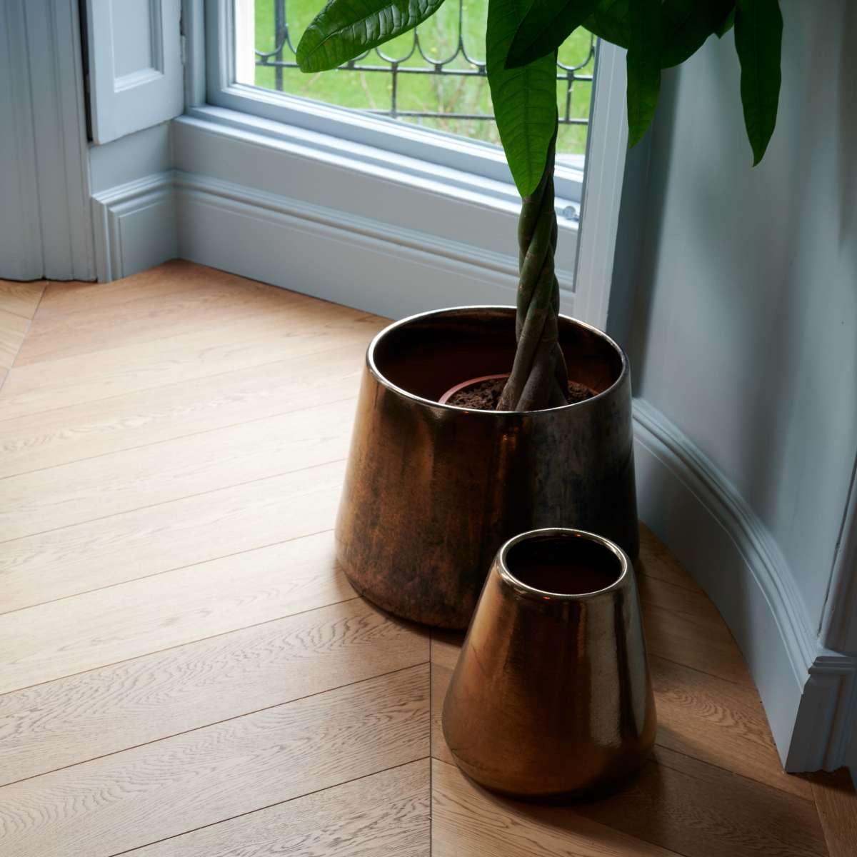A large and a small metal planter are positioned on a wooden floor, near a window.