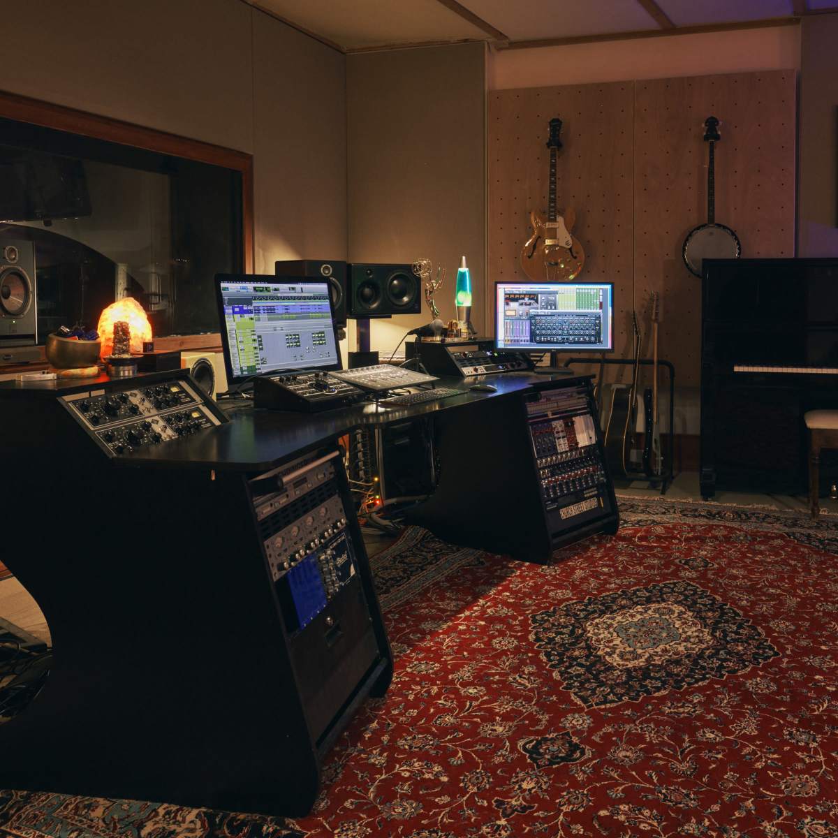 A music studio equipped with audio equipment, computers, and a grand piano, all arranged on a patterned rug.