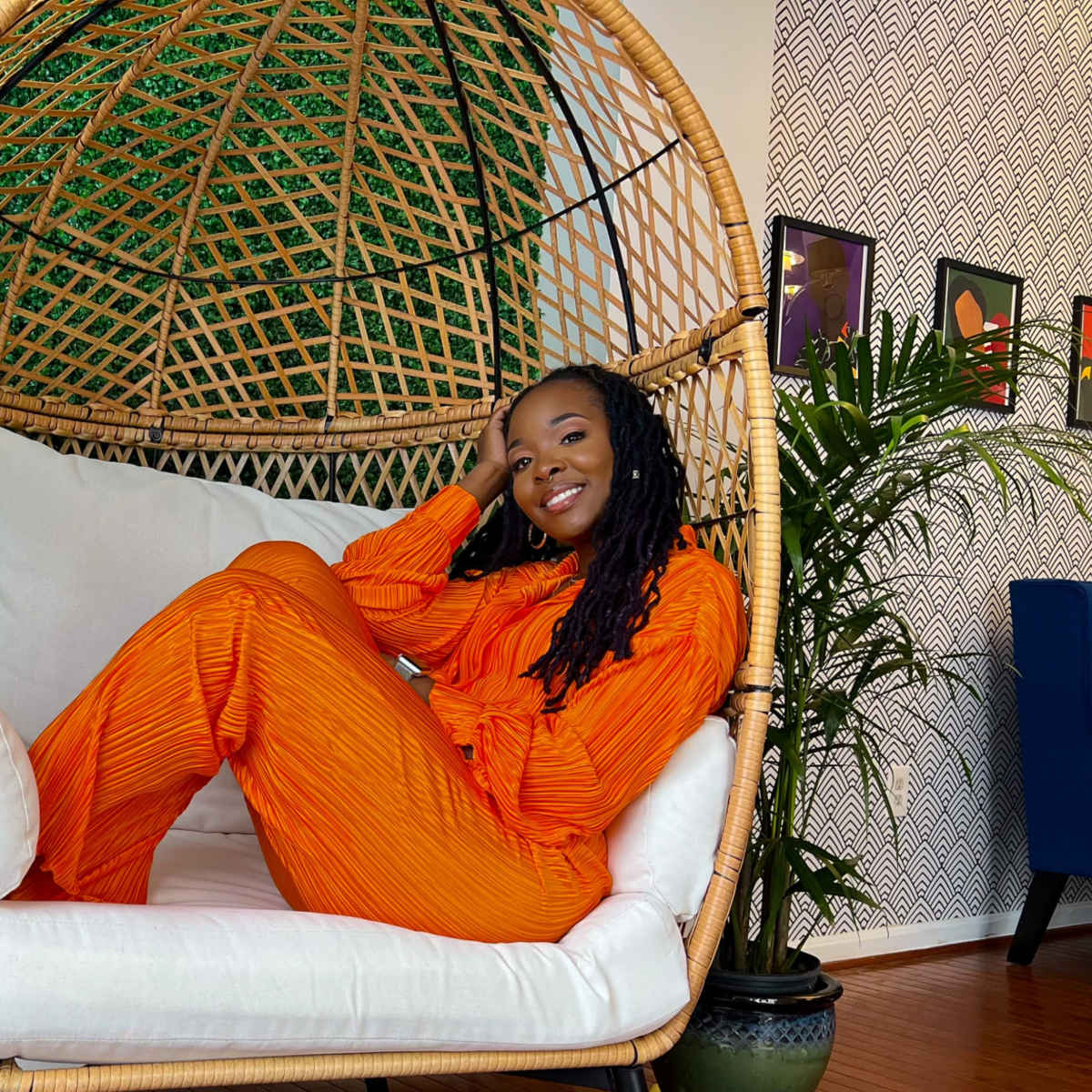 A person wearing an orange outfit sits in a large woven hanging chair surrounded by greenery and indoor plants.
