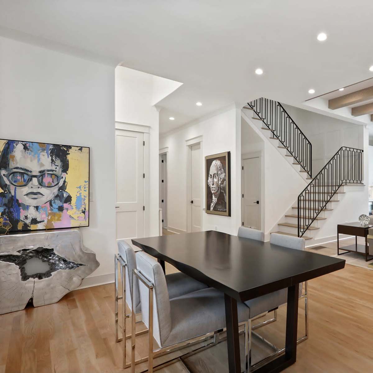 A modern living area with an open floor plan, featuring a dining table, chairs, artwork on the walls, and a staircase leading to an upper level.