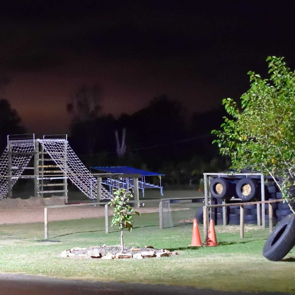 A playground at night featuring a large climbing structure, a sand area, and surrounding trees.