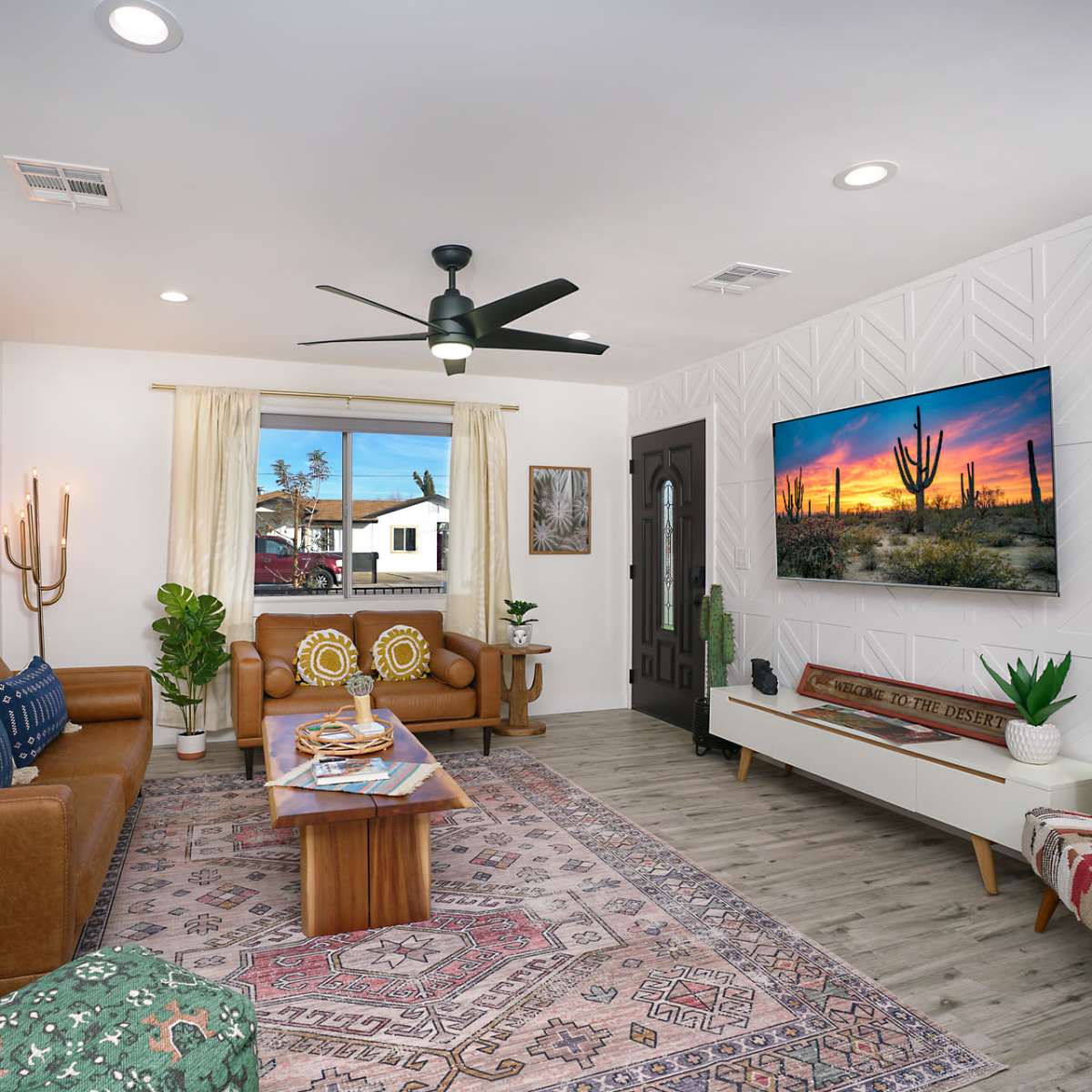 A living room features brown leather sofas, a wooden coffee table, a decorative rug, and a wall-mounted television displaying a desert landscape.