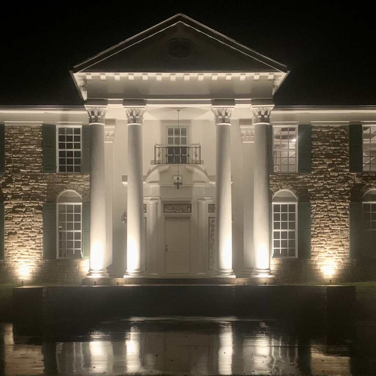 A large, stately mansion is illuminated at night, with prominent architectural features and a reflecting pond in front.