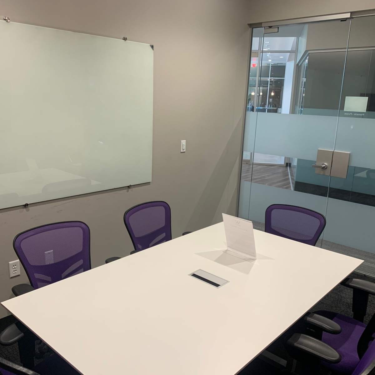 A conference room features a whiteboard, a rectangular table with a central device, and four purple chairs around it.