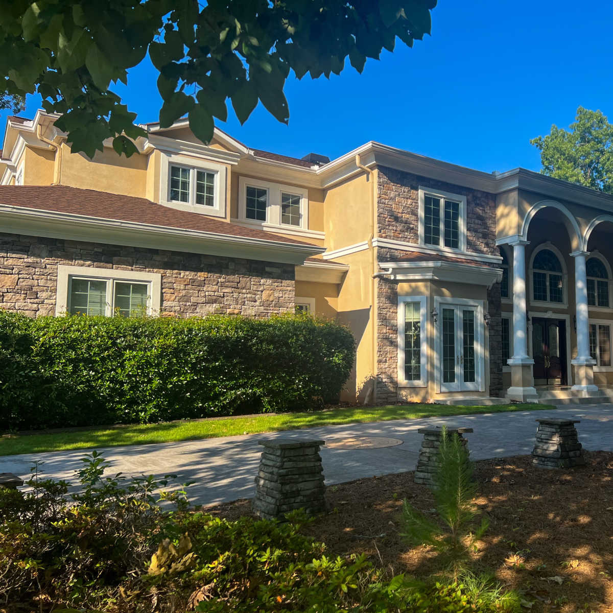 A large, two-story house with a stone and stucco facade, featuring arched entrances and a wide driveway surrounded by landscaping.