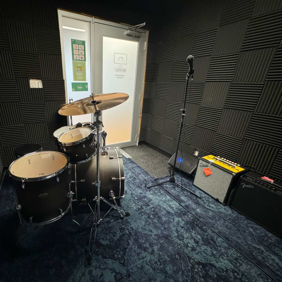 A soundproofed music practice room featuring a drum set, microphone stand, amplifier, and a carpeted floor.