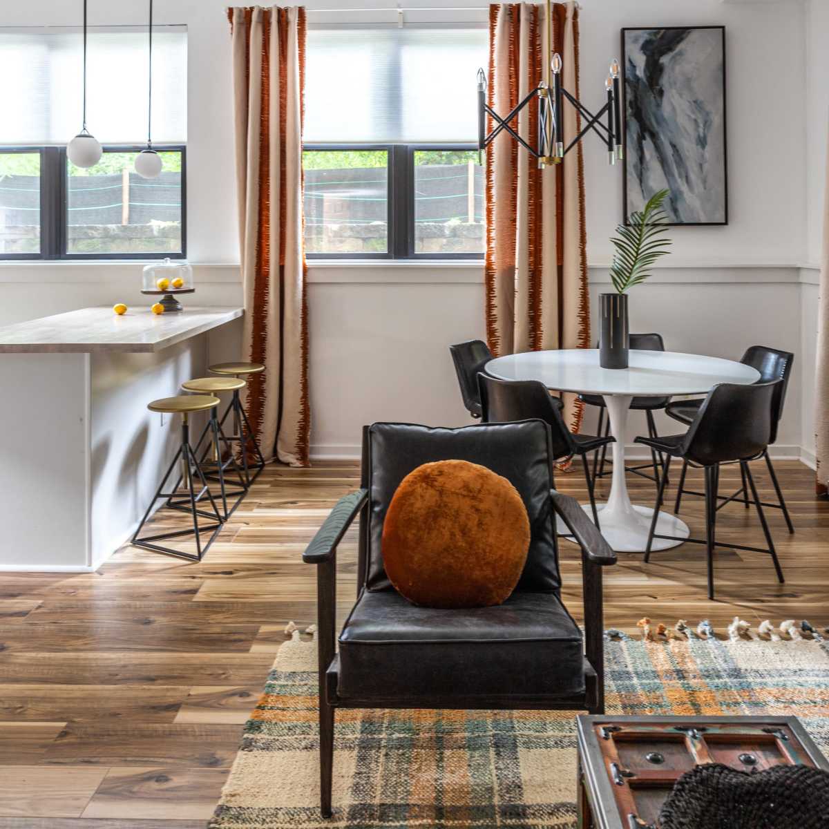The scene depicts a modern living space featuring a black chair with an orange cushion, a round dining table surrounded by chairs, and large windows dressed with striped curtains.