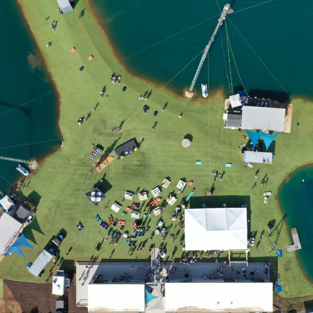 An aerial view of a water sports facility with people gathered on green grass and several water bodies surrounding the area.