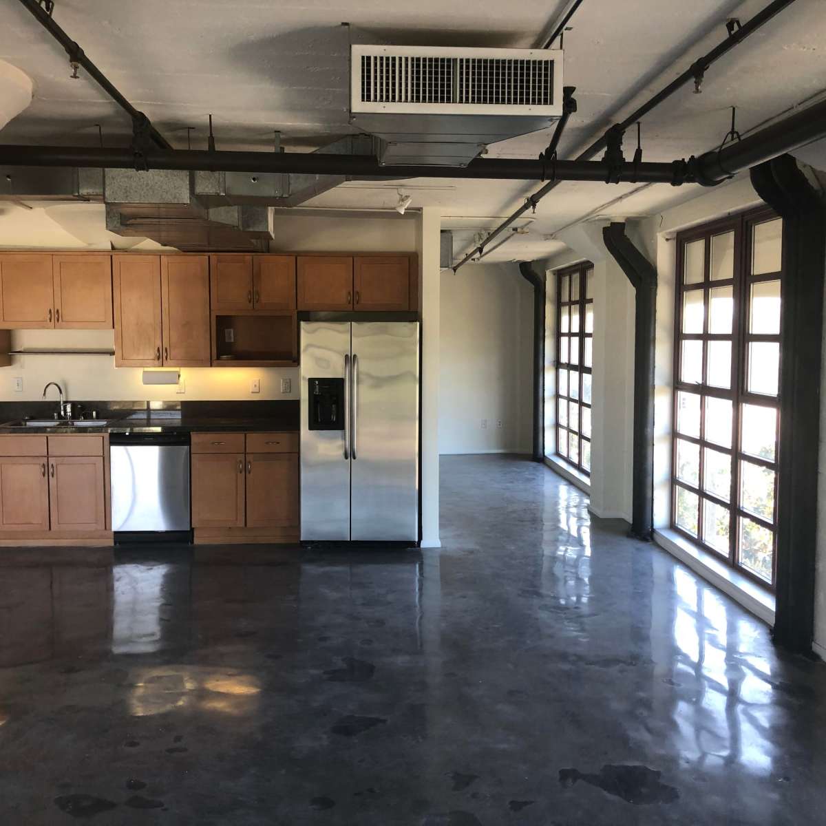 A modern open-concept living space with a kitchen area featuring wooden cabinets and stainless steel appliances, and large windows allowing natural light to enter.