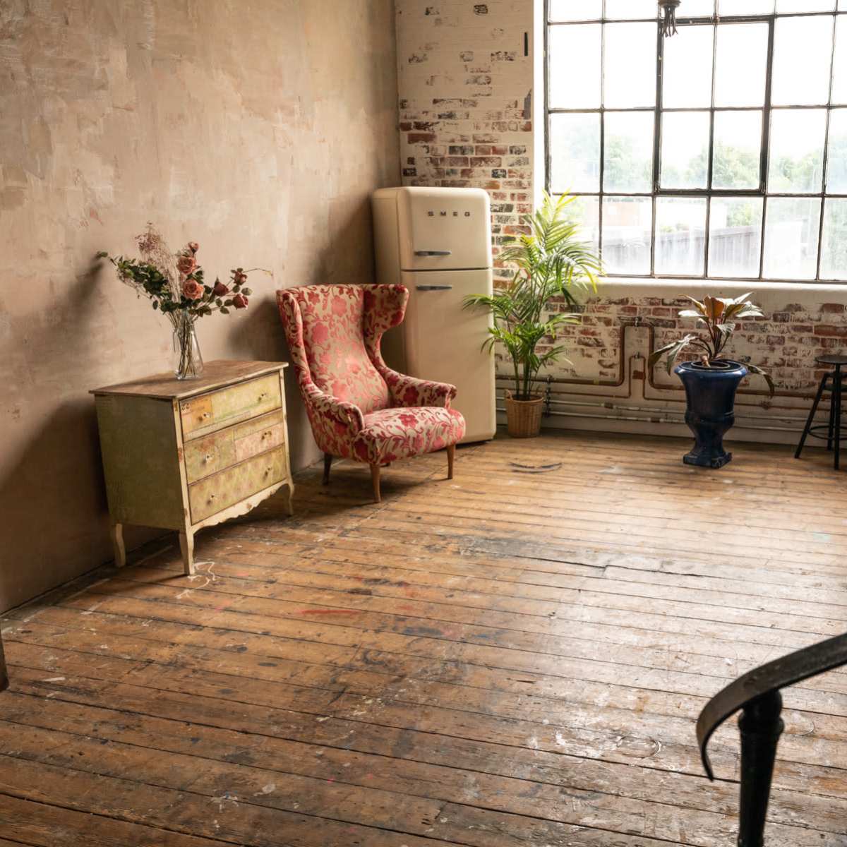 The room features a mix of vintage furniture, including two chairs, a chest of drawers, and a blue planter, with large windows offering a view outside.