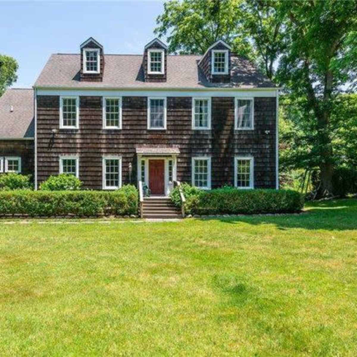A two-story wooden house with a front porch, surrounded by a well-maintained lawn and dense greenery.