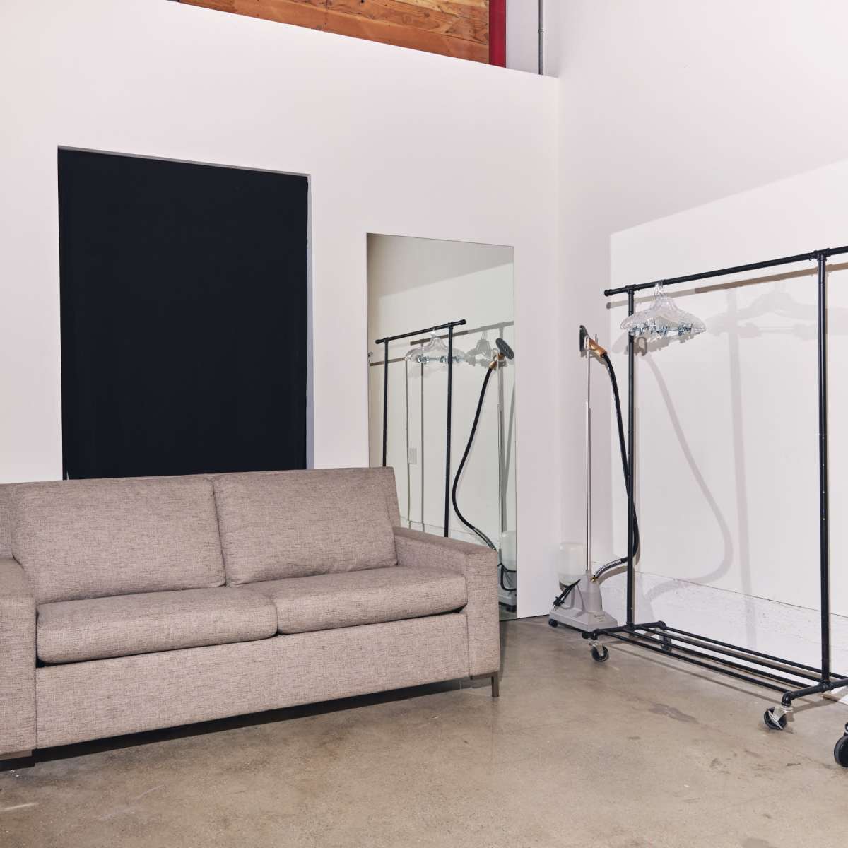A beige sofa situated against a white wall with a black window and a mirror, alongside clothing racks on wheels.