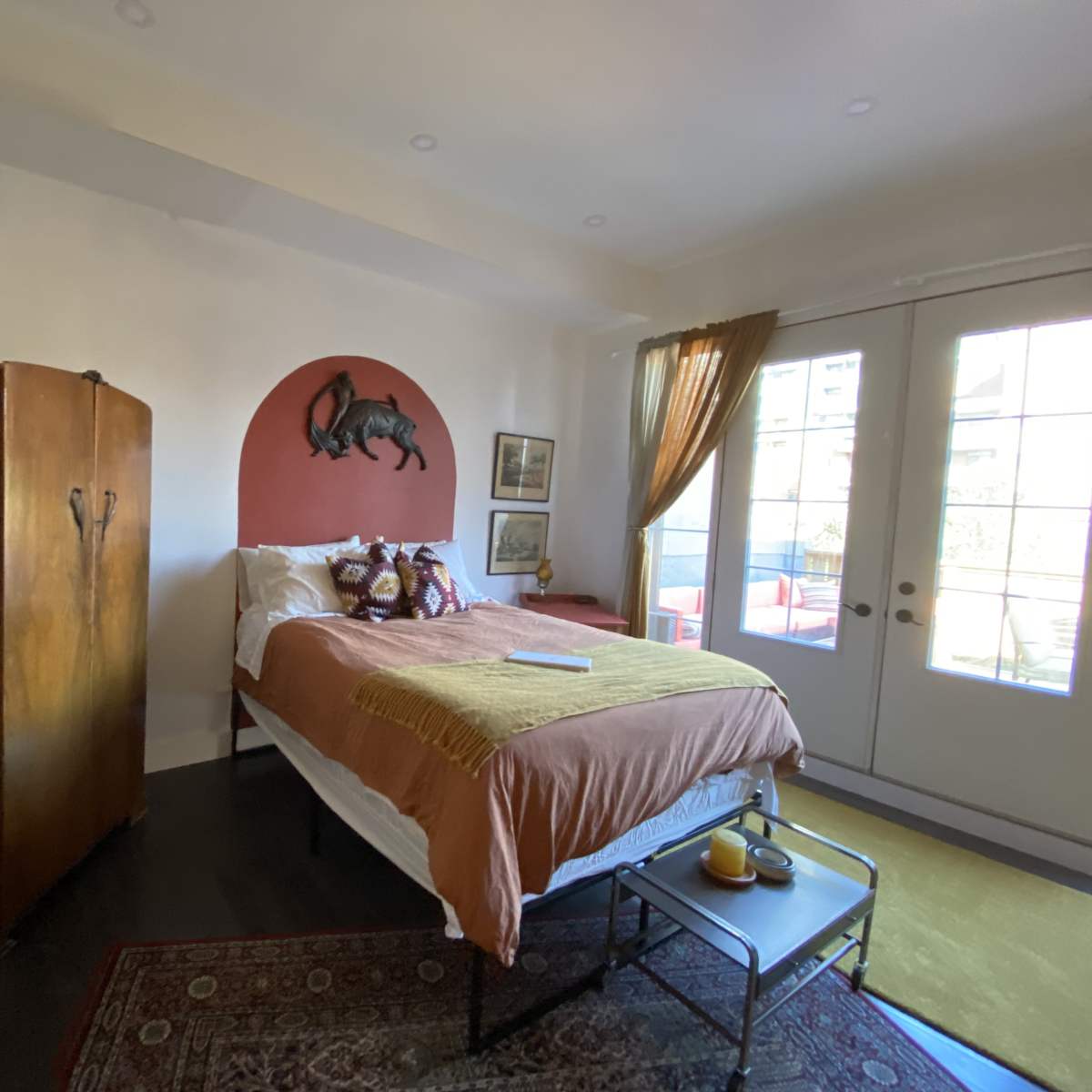 A bedroom features a bed with orange bedding, a wooden wardrobe, and a decorative wall with a horse painting, along with large glass doors opening to an outdoor area.
