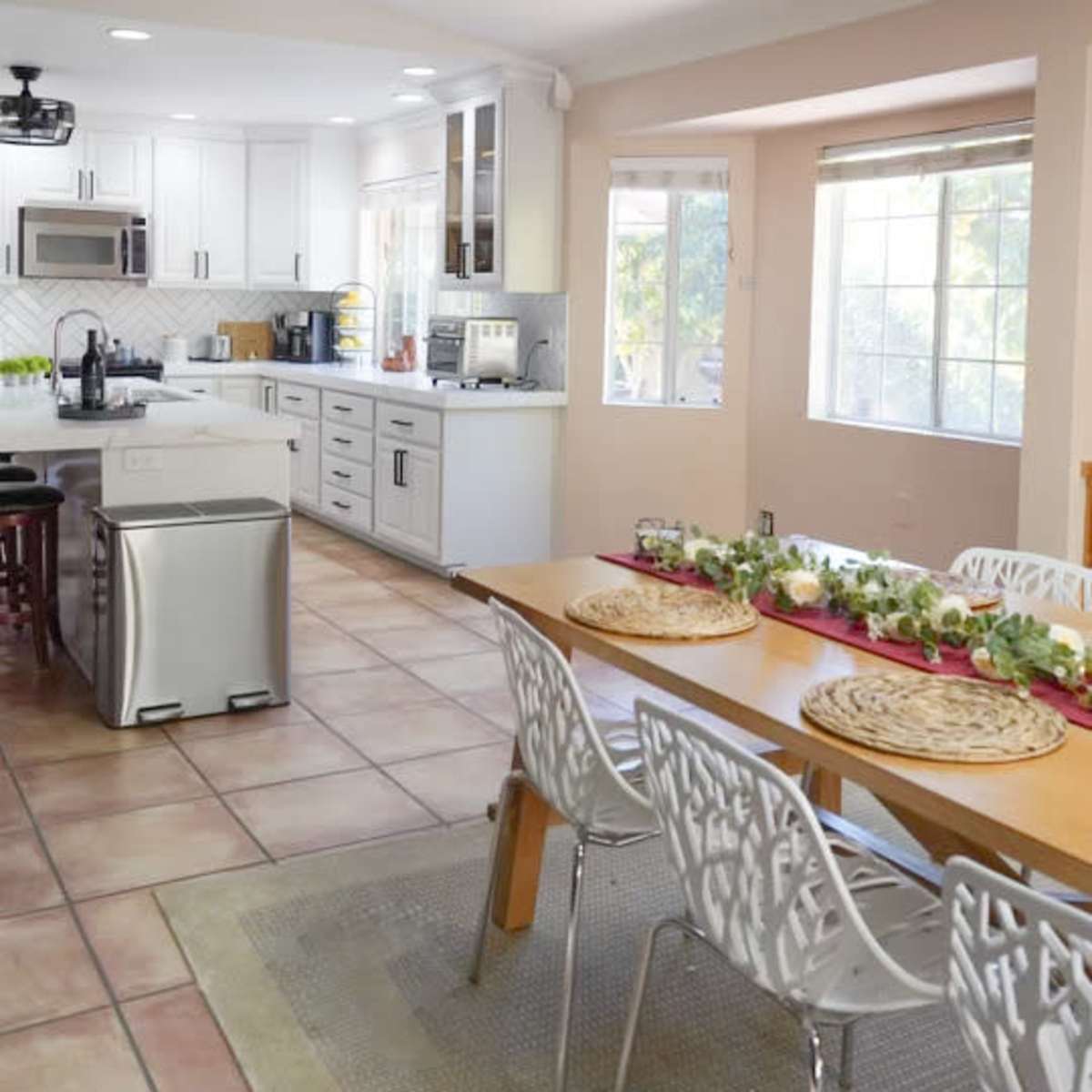 A modern kitchen area features a dining table with white chairs, a large window allowing natural light, and an organized layout with appliances and cabinetry.