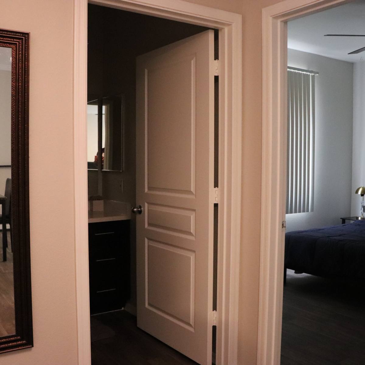 A hallway leading to two separate rooms, with a mirror on the left and a view into a dining area and a bedroom.