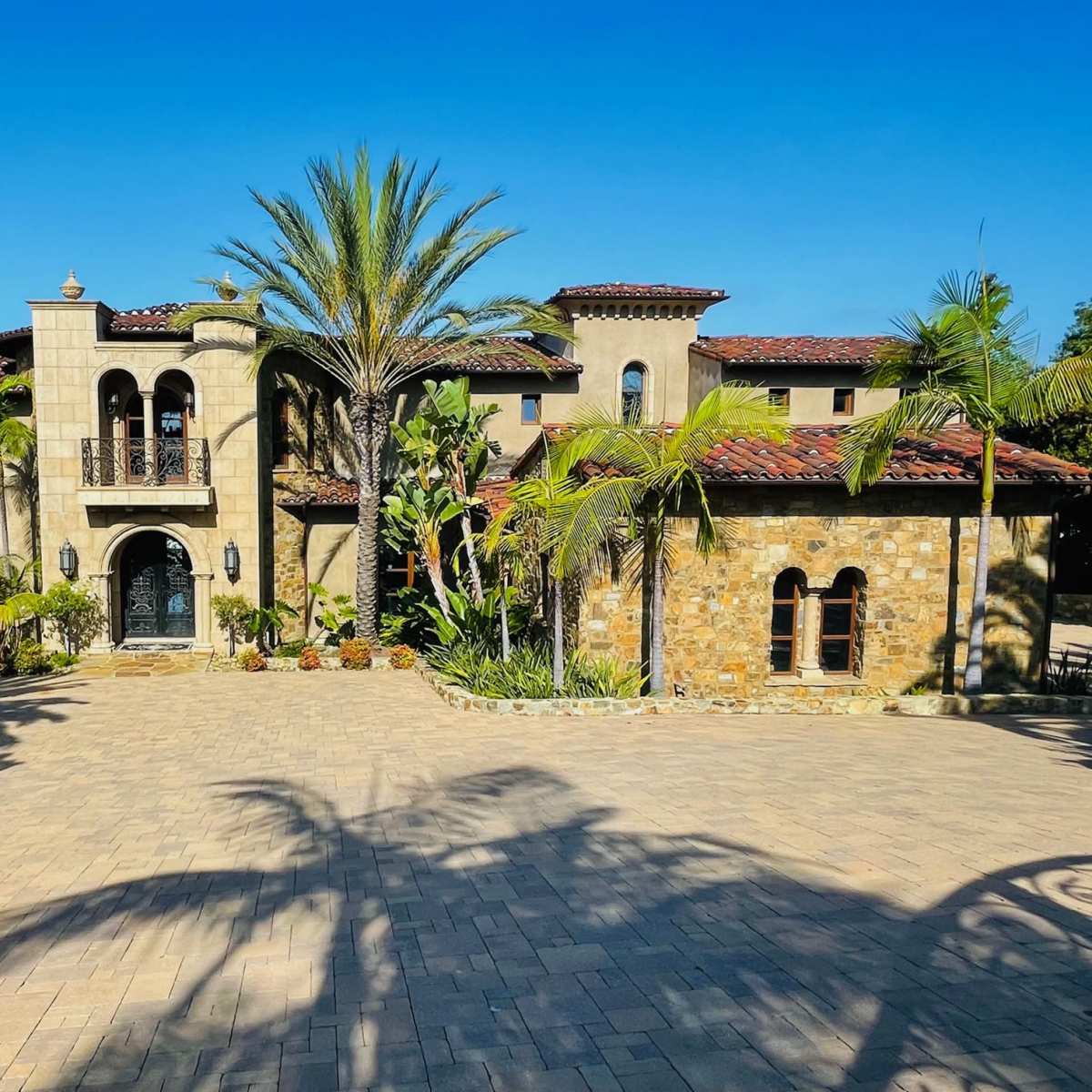 A large Mediterranean-style house featuring stone and stucco exterior with a tiled roof, surrounded by palm trees and a paved driveway.