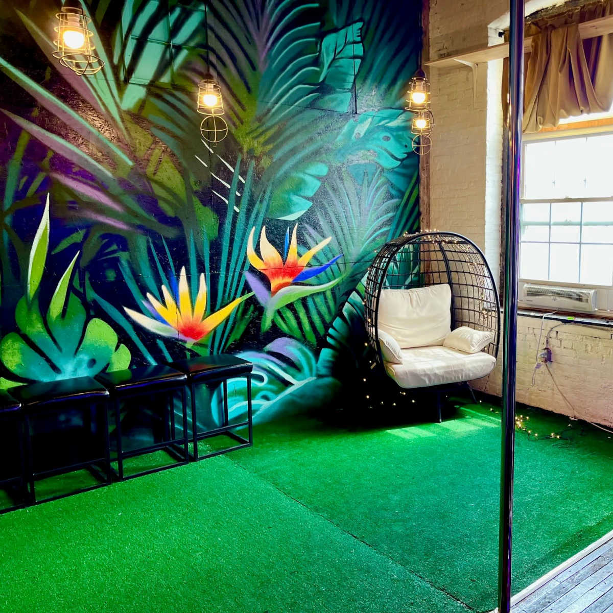 A room with a vibrant tropical mural, featuring a hanging chair, two stools, a television, and a simulated grass floor.