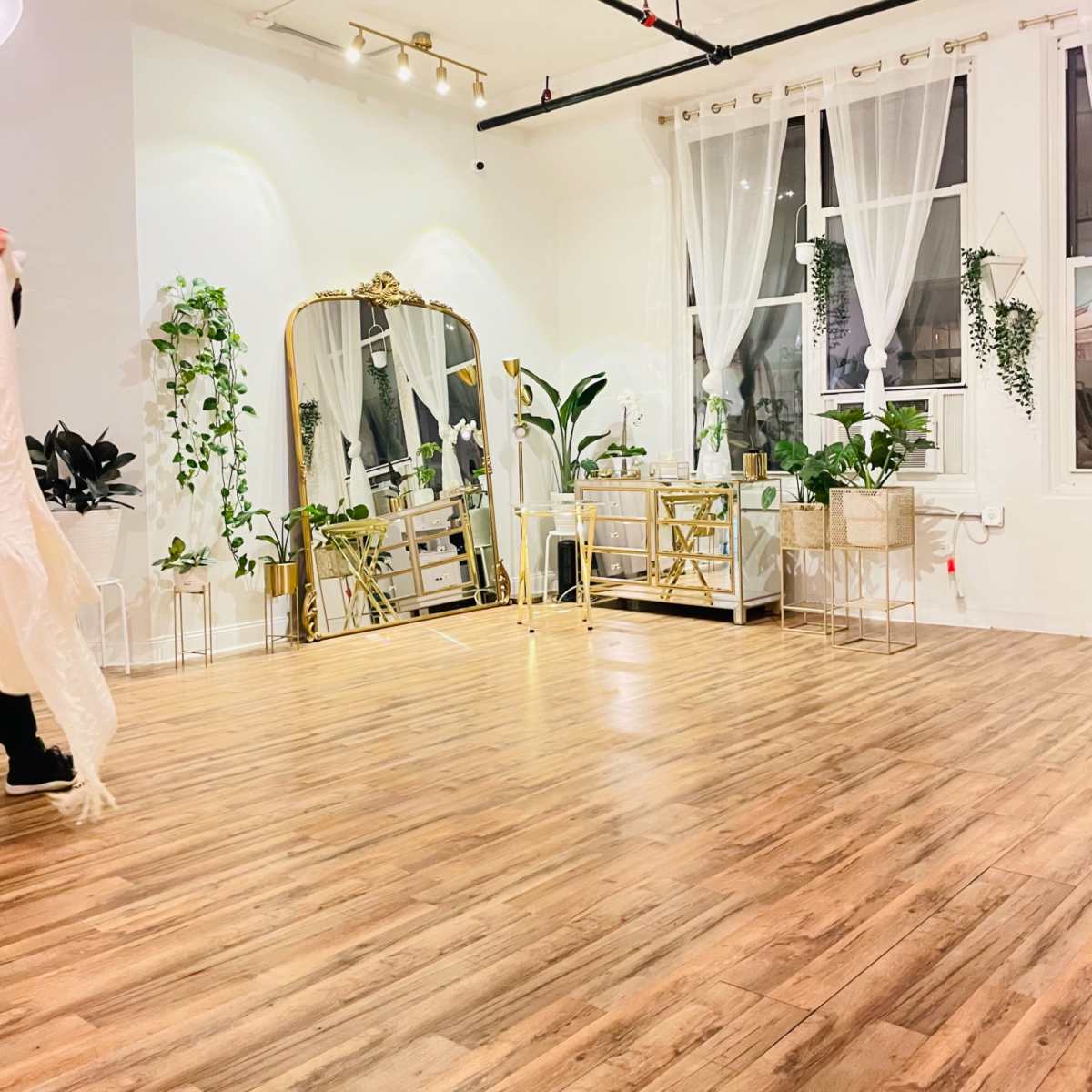 A spacious room features wooden flooring, large windows with sheer curtains, a vintage mirror, and various indoor plants arranged around a console table.