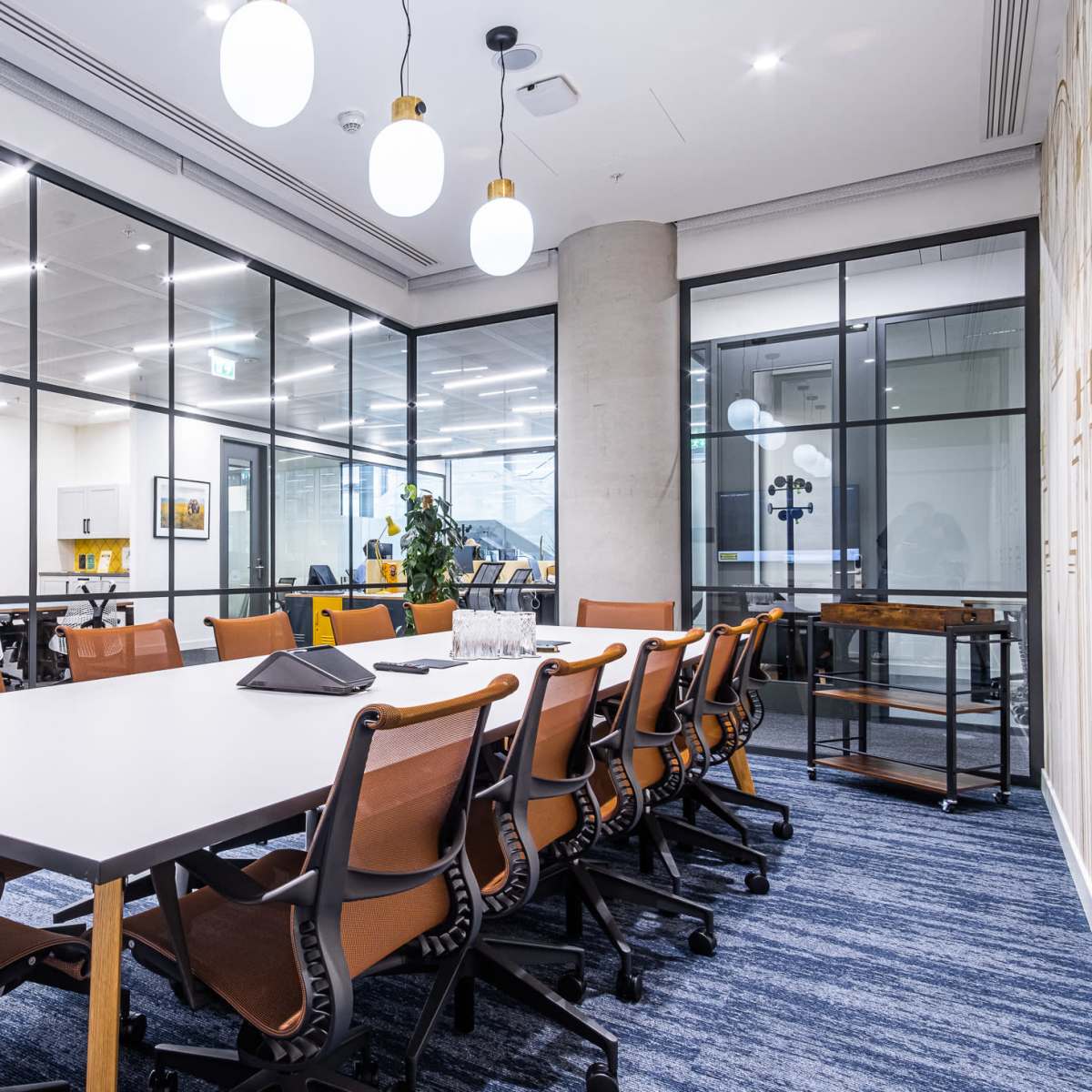 A modern office meeting room with a large table surrounded by chairs, glass walls, and a few employees working at desks in the background.