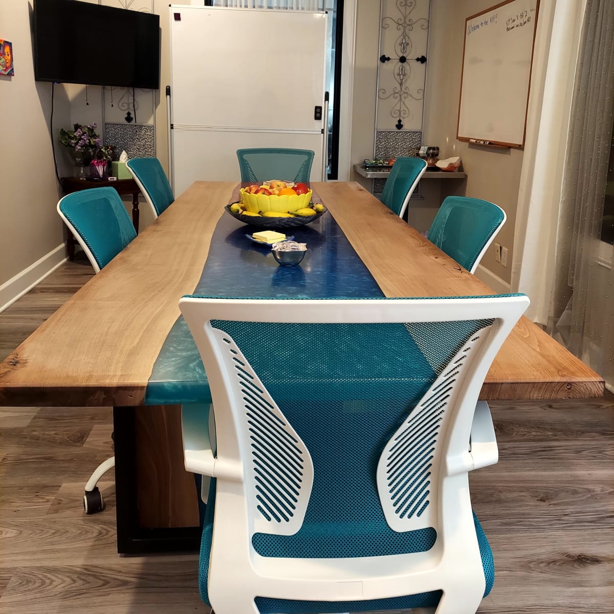 A conference room features a long wooden table with turquoise chairs, a fruit bowl at the center, and a television on one wall.