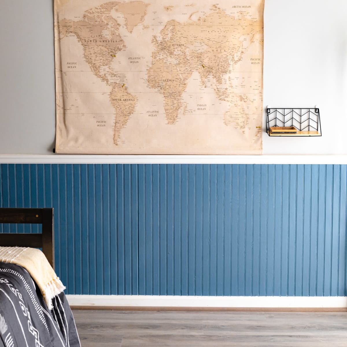 A world map hanging on a wall above a blue wainscoted section in a room with wooden flooring and a bed partially visible.