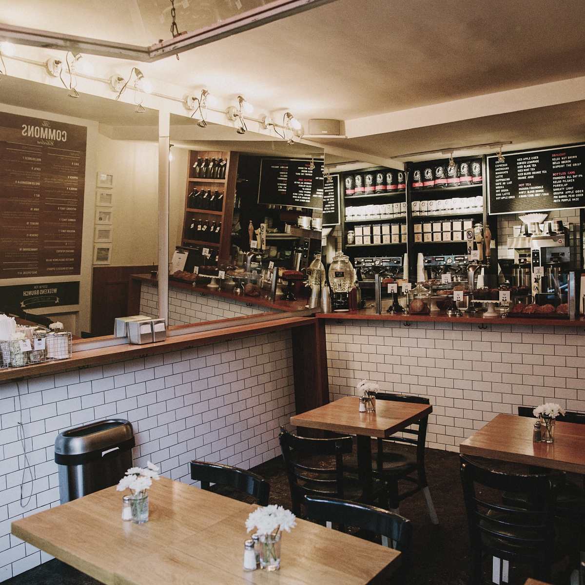 A cozy cafe interior with wooden tables, a barista counter displaying coffee machines, and a wall of shelves filled with various beverages.