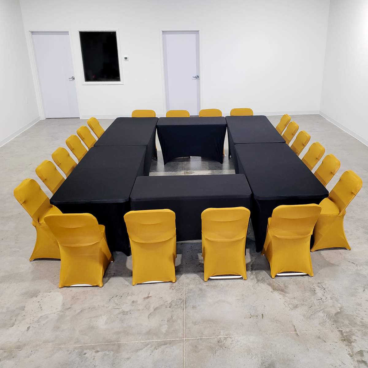 A meeting room features an arrangement of black tables in a square shape surrounded by yellow-covered chairs.