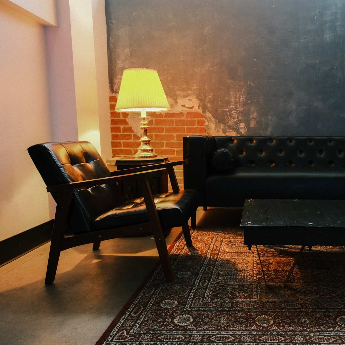 A black tufted sofa and a wooden armchair are arranged around a low coffee table on a patterned rug in a room with exposed brick walls.