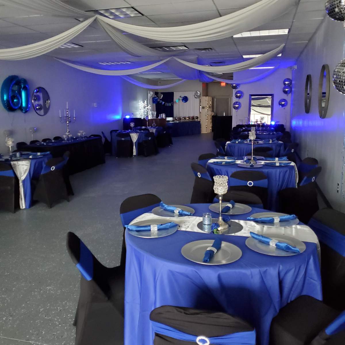 A decorated event space with tables covered in blue tablecloths and black chairs, set up for a celebration with blue and silver accents.