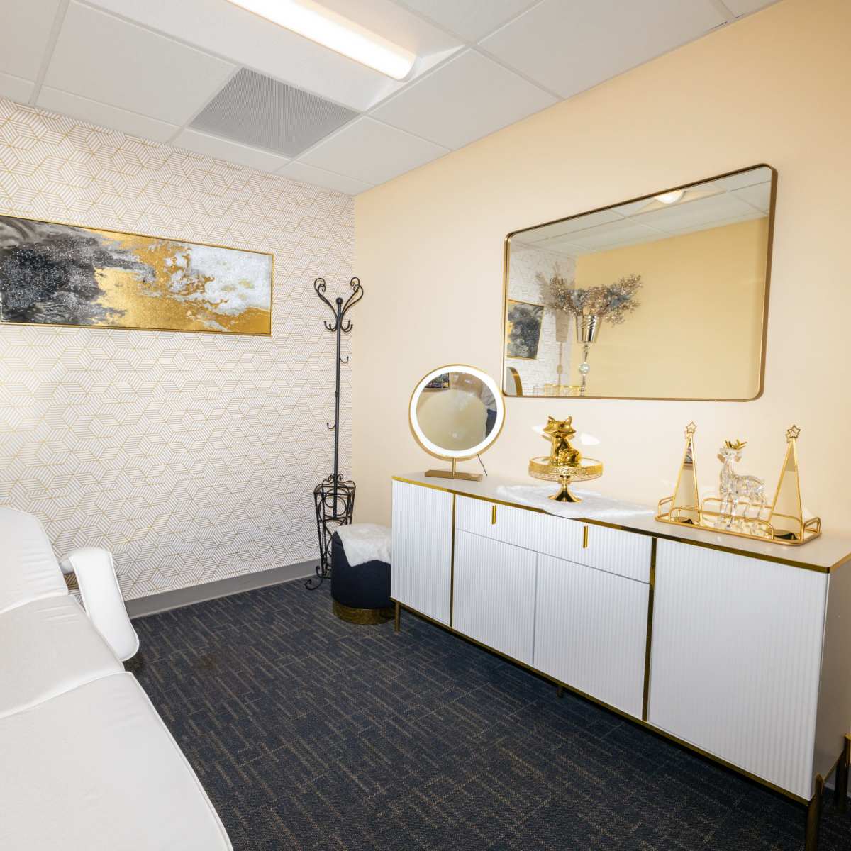 A minimalist treatment room featuring a massage table, a decorative mirror, a small cabinet, and a wall adorned with geometric art.