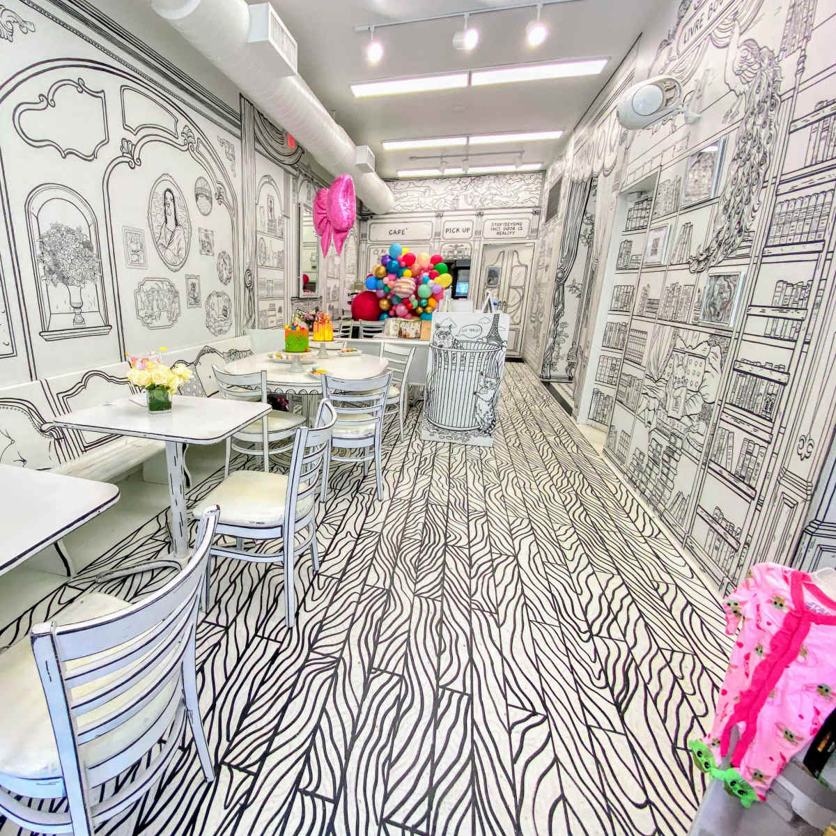 A brightly lit café with black and white illustrated walls, floor, and furniture, featuring tables and chairs arranged neatly with floral decorations.