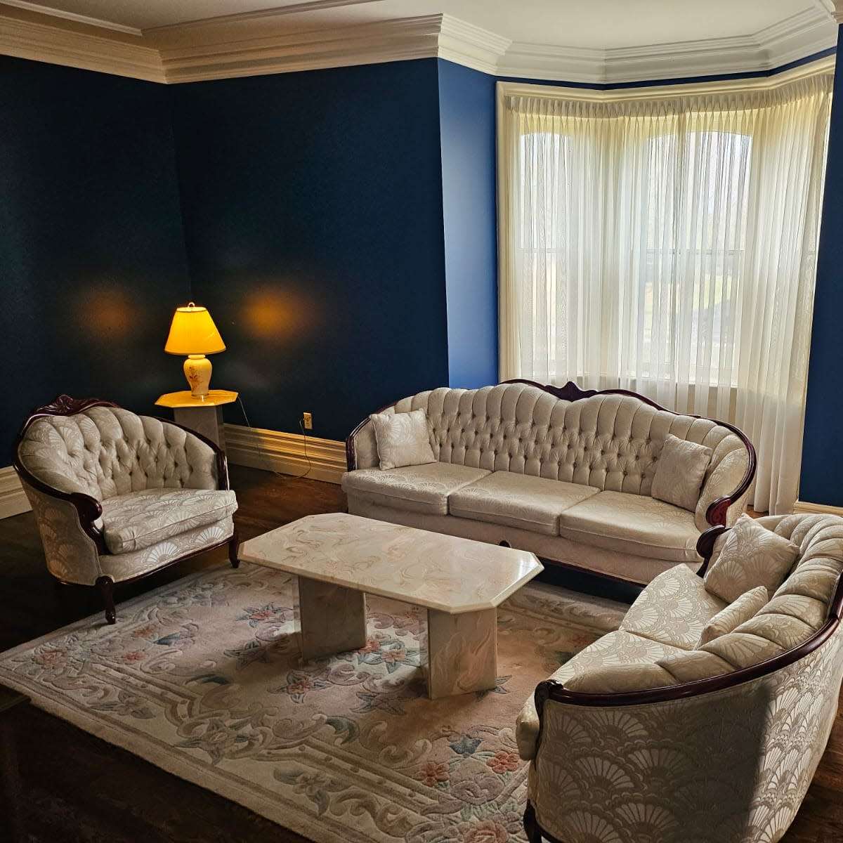 A beautifully furnished living room with a light-colored upholstery sofa set, a marble coffee table, and two lamps illuminating the space against a dark blue wall.